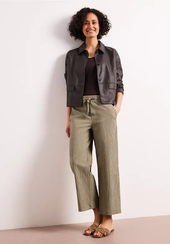 Vrouw met krullend haar draagt een zwarte trui, donker jasje, olijfgroene wijde broek en sandalen voor een lichte achtergrond.