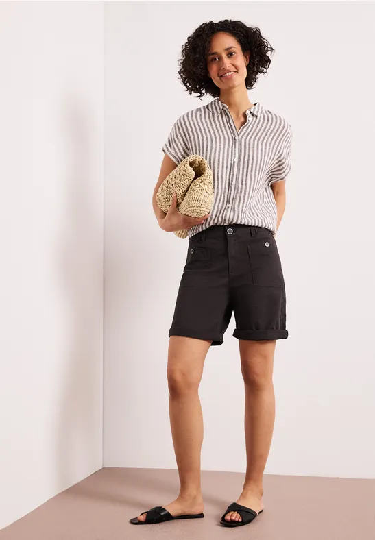 Persoon in een hoek van de kamer draagt gestreept overhemd, zwarte shorts en sandalen, met strooien tas.