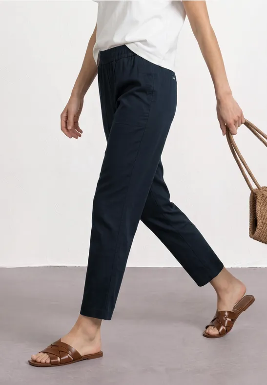Vrouw in wit shirt, donkerblauwe 7/8-broek, platte sandalen en met een rieten tas in de hand.