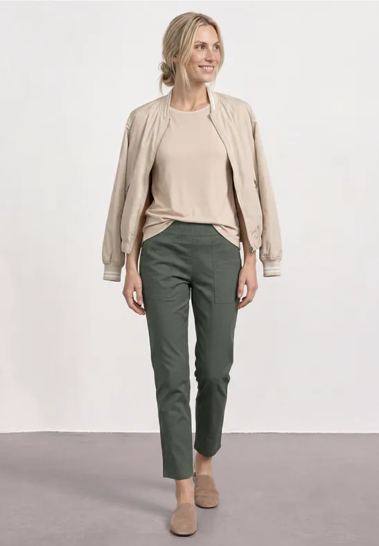 Eine Frau trägt eine beige Jacke, ein beiges Shirt, dunkelgrüne Hose und beige Schuhe vor neutralem Hintergrund.