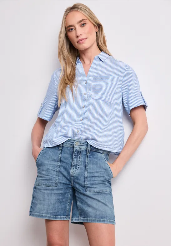 Eine Frau trägt ein hellblaues Hemd und Jeans-Shorts, die Hände in den Taschen