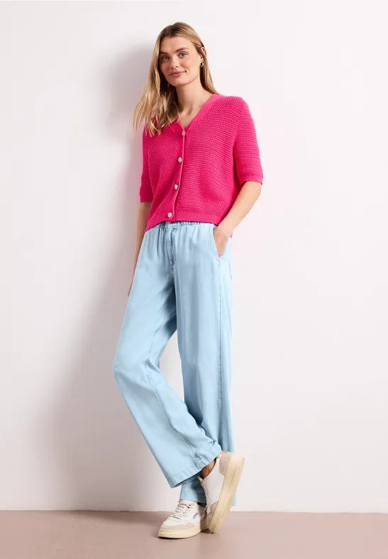 Vrouw in roze vest, lichtblauwe wijde broek en witte sneakers, staand ontspannen tegen lichte achtergrond.