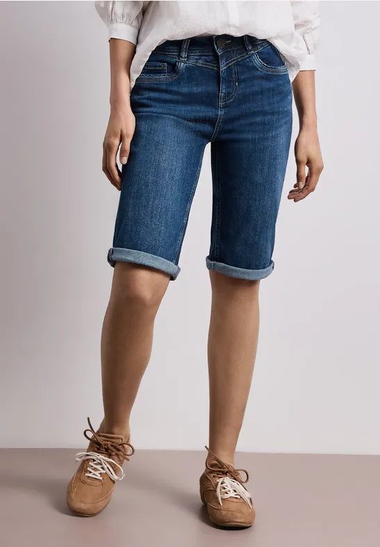 Beinansicht mit blauem Jeans-Bermuda, weißem Oberteil und hellbraunen Wildlederschuhen.