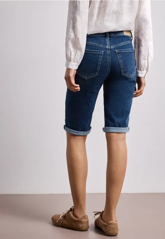 Frau von hinten in blauer Jeans-Bermuda, weißer Bluse und braunen Loafers.