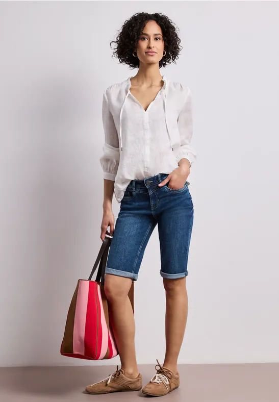 Frau in weißer Bluse, Jeansbermuda und Sandalen hält pink-rote Tasche.