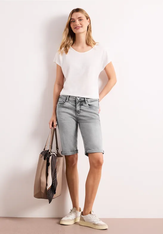 Vrouw in wit t-shirt, grijze denim bermuda en witte sneakers, draagt een grote tas.