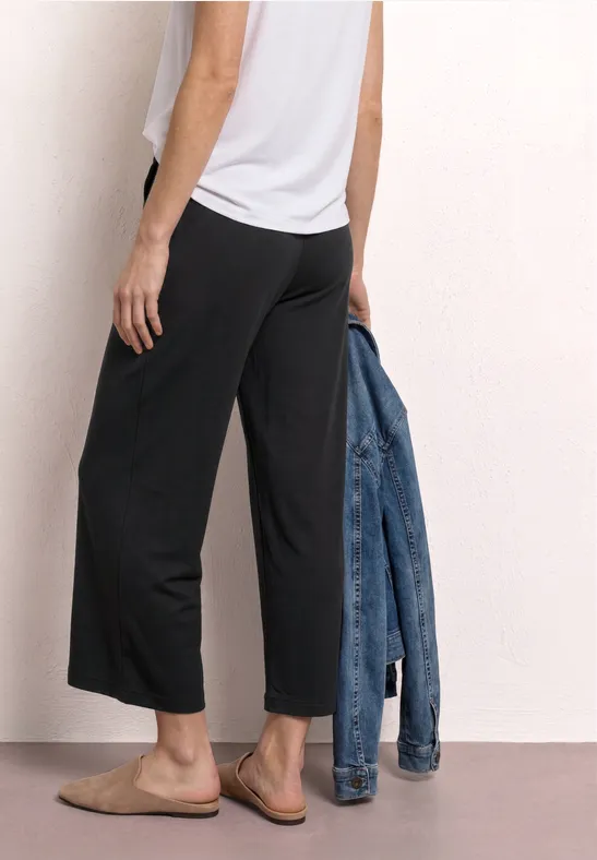 Frau trägt weißes Top und schwarze Culotte-Hose, hält Jeansjacke und trägt Slipper.