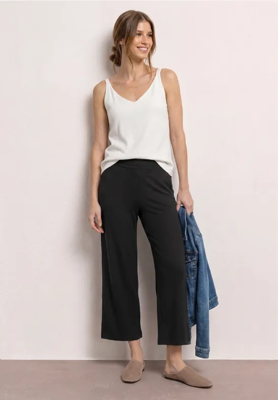 Frau in weißem Top, schwarzen Culottes und taupe Mules, mit Jeansjacke in der Hand.