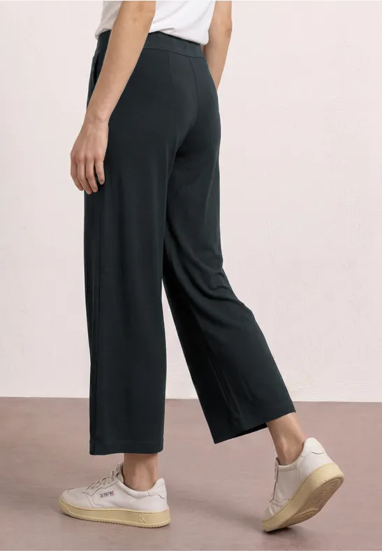 Eine Person trägt schwarze, weit geschnittene, knöchellange Hosen und weiße Sneakers, seitliche Rückansicht.