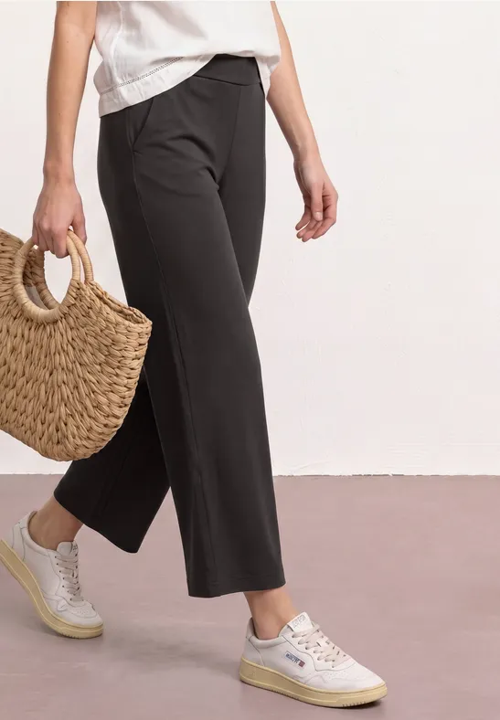 Schlanke 7/8-Hose in Schwarz, weiße Sneakers und Basttasche auf hellem Boden