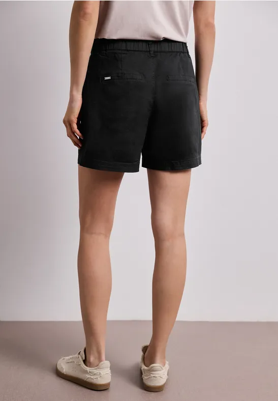 Person von hinten in schwarzen Shorts, weiße Sneakers, neutrale Wand im Hintergrund