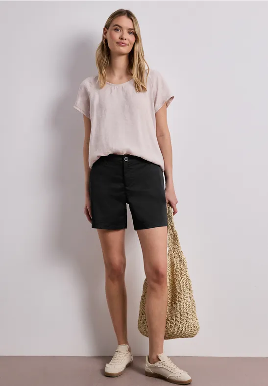 Frau in weißem T-Shirt, schwarzen Shorts, beigen Sneakern und Strohtasche vor hellem Hintergrund.