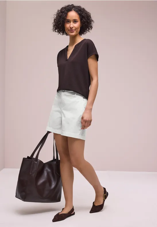 Frau mit lockigem Haar in dunklem T-Shirt, hellem Rock, schwarzen Ballerinas und großer brauner Tasche.