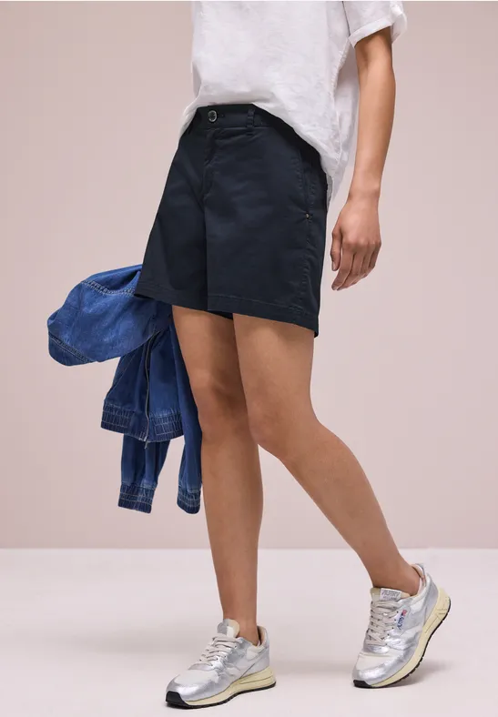 Eine Person trägt dunkelblaue Shorts, weißes T-Shirt, hält eine Jeansjacke und trägt silberne Sneaker.