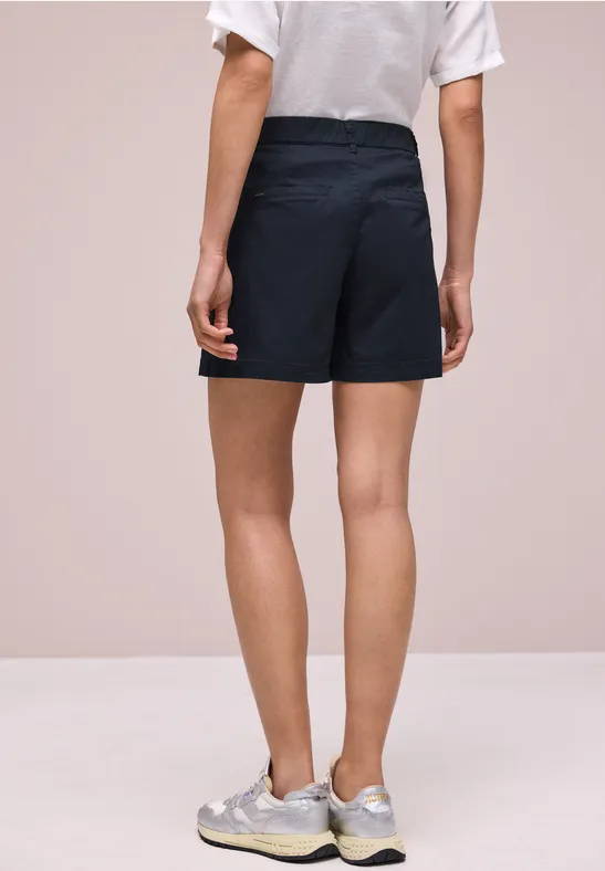 Frau von hinten in dunkelblauen Shorts, weißem T-Shirt und Sneakern vor hellem Hintergrund.