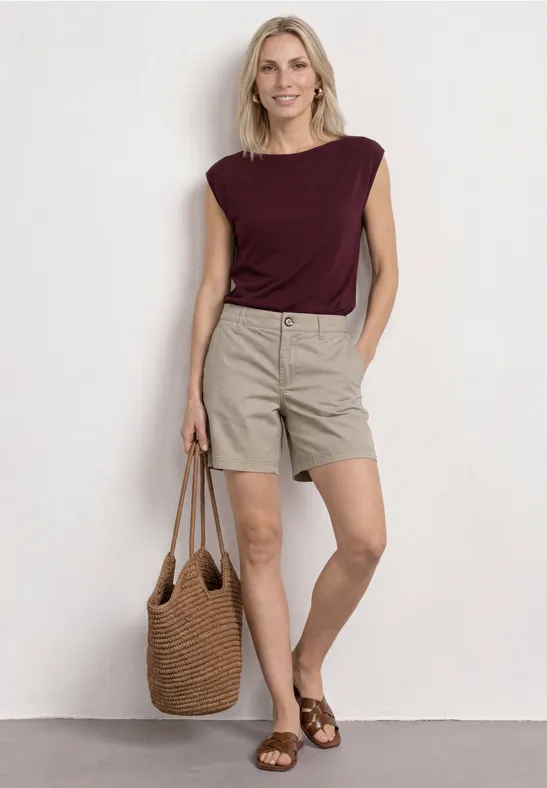 Vrouw in bordeaux top, beige shorts en bruine sandalen met strozak, staand voor een lichte achtergrond.