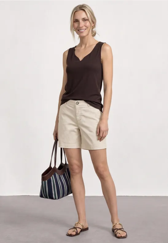 Vrouw in bruin hemd en beige short, sandalen en gestreepte tas, voor een witte achtergrond.