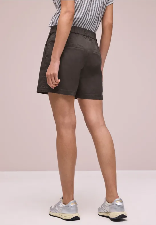 Rückansicht einer Person in schwarzen Shorts und hellen Sneakers vor neutralem Hintergrund.