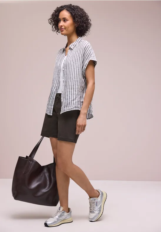Frau mit lockigen Haaren trägt gestreifte Bluse, schwarze Shorts, Sneaker und große braune Tasche.