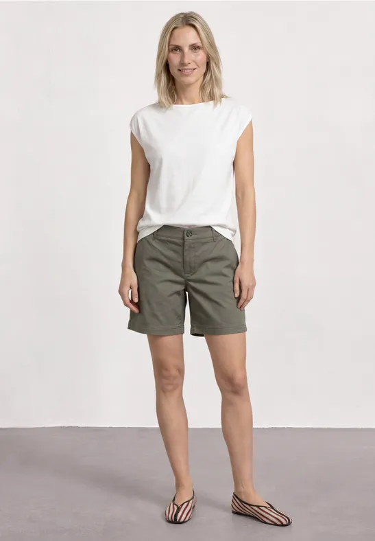 Eine Frau in weißem T-Shirt, khakigrünen Shorts und gestreiften Pantoletten vor neutralem Hintergrund.