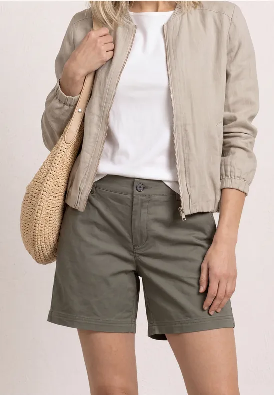 Frau trägt khakifarbene Shorts, weißes T-Shirt, beigen Blouson und Basttasche vor hellem Hintergrund.