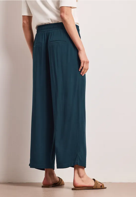 Persoon van achteren in losse, petrolblauwe broek tot de enkels en sandalen.