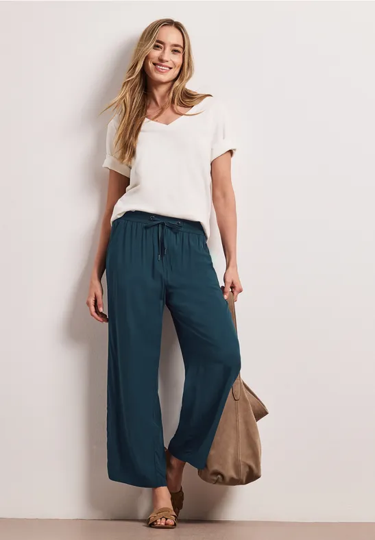 Vrouw in wit T-shirt en losse turkooizen broek, sandalen aan de voeten en draagt een tas