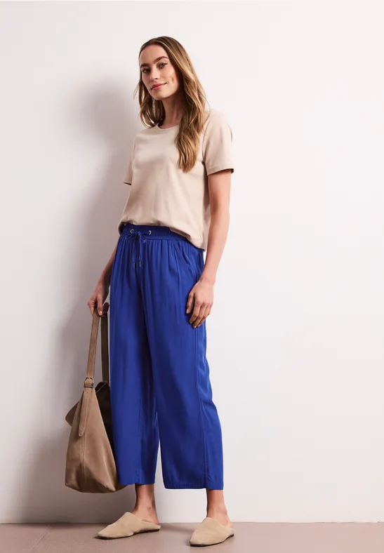 Vrouw in een wit casual t-shirt, blauwe wijde broek, beige slip-ons en een grote bruine tas.