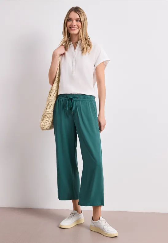 Person im weißen Shirt, weiten grünen Culottes, hellen Sneakern und Korbtasche.