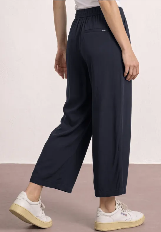 Vrouw draagt wijde donkerblauwe broek met elastische taille tot op de enkel en witte sneakers.