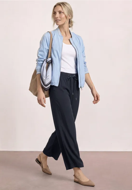 Vrouw met lichtblauwe jas, wit topje, donkere broek en nude ballerinas, met twee tassen.