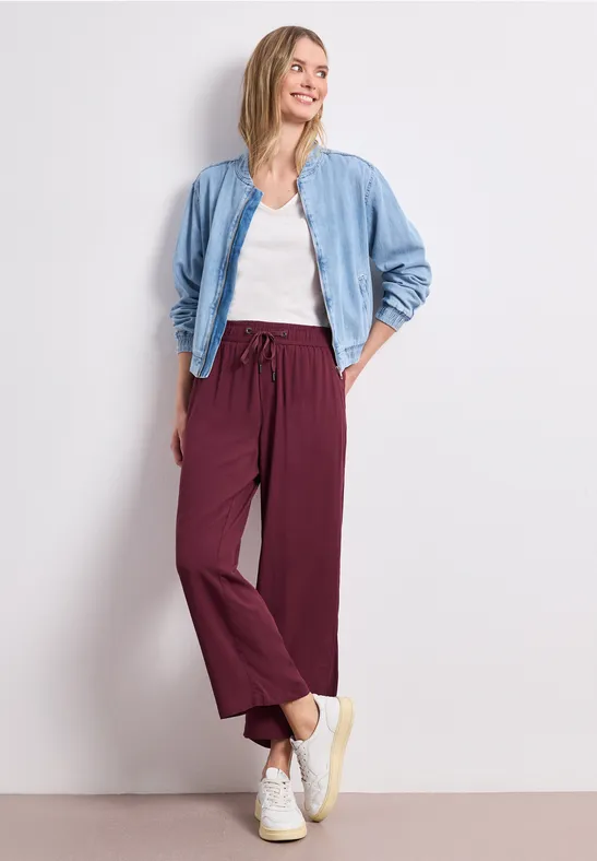 Frau lehnt an weißer Wand und trägt weißes Shirt, helle Jeansjacke, weinrote weite Hose und weiße Sneaker.