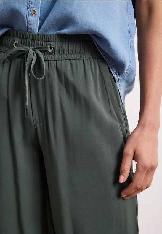 Persoon draagt een olijfgroene broek met trekkoord en een lichtblauw overhemd, rechterhand naast het lichaam.