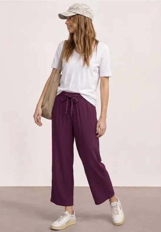 Vrouw in wit T-shirt, paarse wijde broek, sneakers en pet voor een neutrale achtergrond.