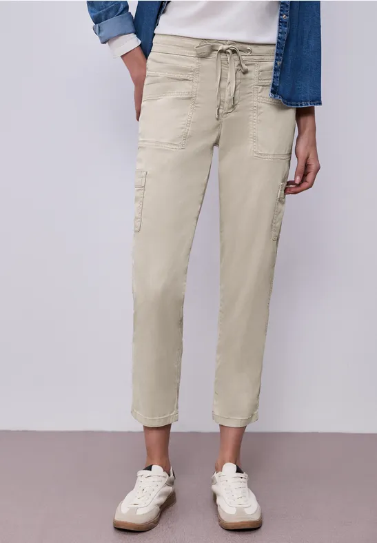 Beige Cargo-Hose mit Tunnelzug, seitlichen Taschen und weißen Sneakern an neutraler Wand.