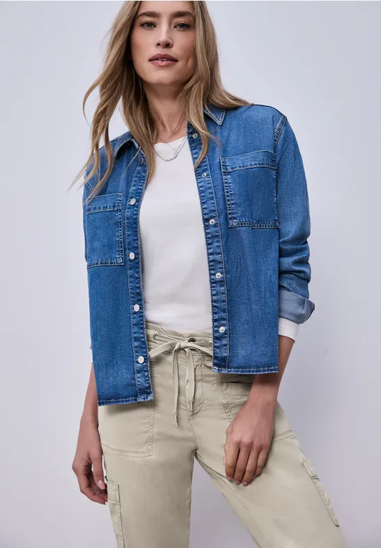 Person steht vor hellem Hintergrund in blauer Jeansjacke, weißem Shirt und beigen Hosen.