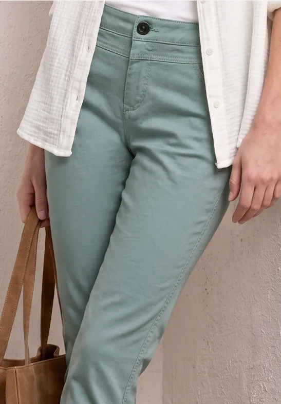 Draagt een witte blouse, een open textuurovershirt, mintgroene chino en houdt een bruine tas vast.