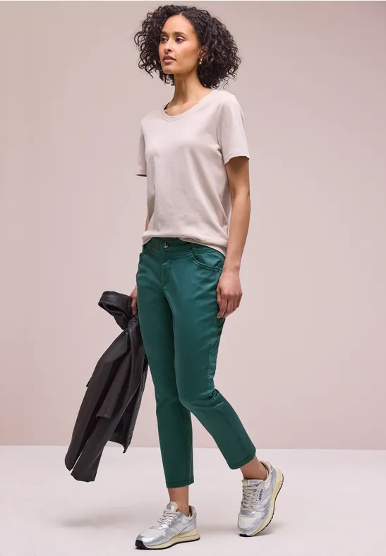 Frau trägt weißes T-Shirt, grüne Hose und Sneaker, hält eine dunkle Jacke.