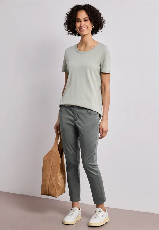 Frau mit lockigen Haaren trägt graues T-Shirt, dunkelgraue Hose, weiße Sneaker und hält eine braune Tasche.