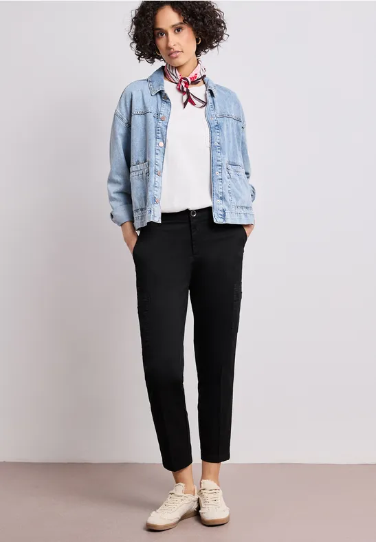 Iemand draagt een spijkerjasje, een wit overhemd, een sjaal, zwarte broek en witte sneakers.