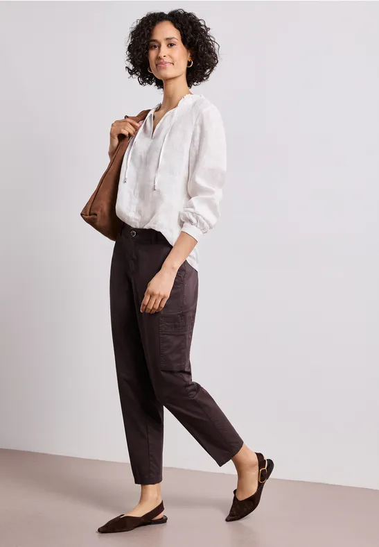 Vrouw met krullend haar draagt witte blouse, donkerbruine broek en loafers, met een bruine tas.