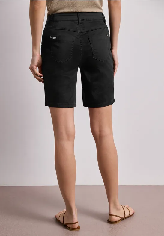 Rückansicht einer Person in schwarzen Bermudashorts und Sandalen auf hellem Boden.