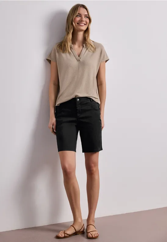 Frau in beigem Oberteil, schwarzen Jeans-Shorts und Sandalen vor schlichtem Hintergrund.