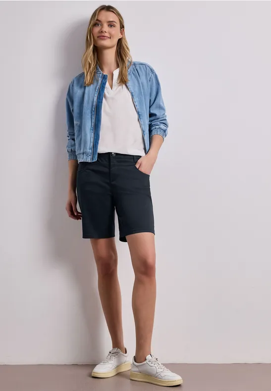 Frau vor weißer Wand trägt Jeansjacke, weißes T-Shirt, schwarze Bermudashorts und weiße Sneaker.