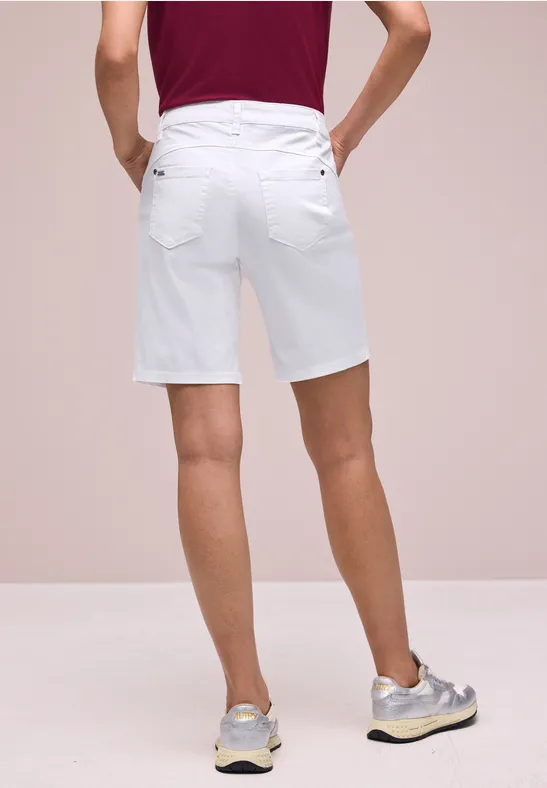 Ein Mann von hinten in weißen Bermudashorts und grauen Sneakern vor neutralem Hintergrund.