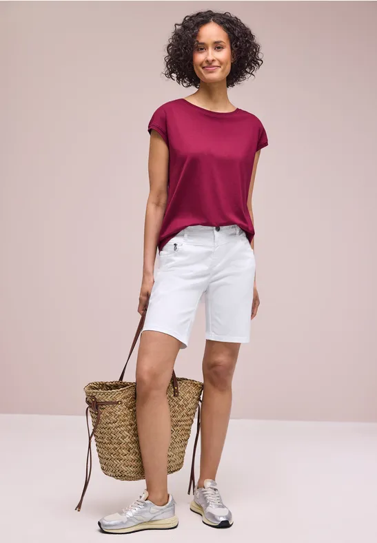 Eine Frau trägt ein weinrotes T-Shirt, weiße Shorts und Sneakers, hält eine geflochtene Strandtasche.