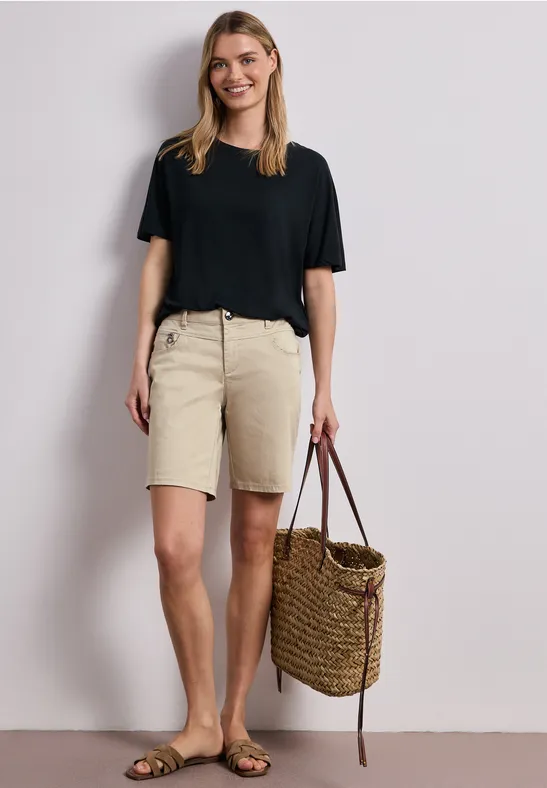 Eine Frau trägt ein schwarzes T-Shirt, beige Bermudashorts, Sandalen und hält eine geflochtene Korbtasche.