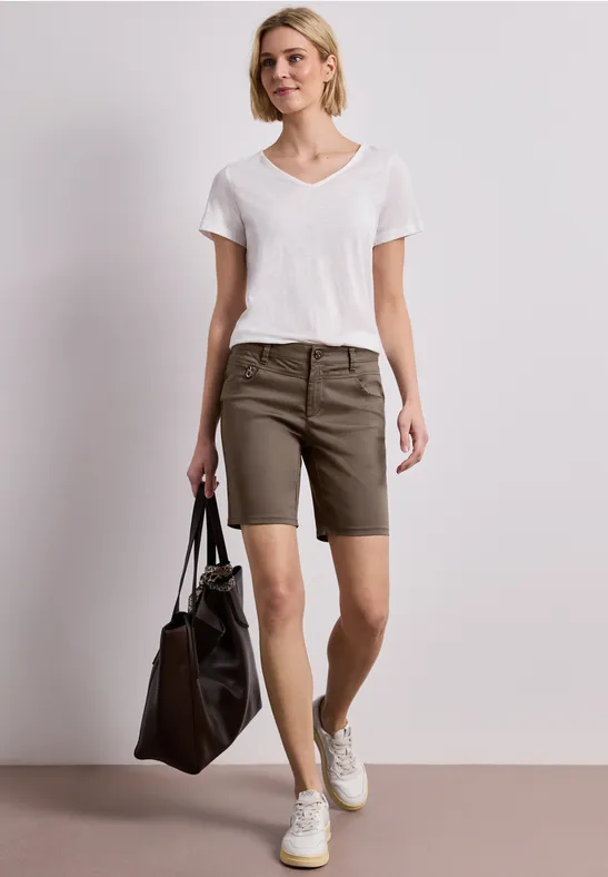 Vrouw in wit T-shirt, kaki korte broek en witte sneakers, met zwarte tas tegen lichte achtergrond.