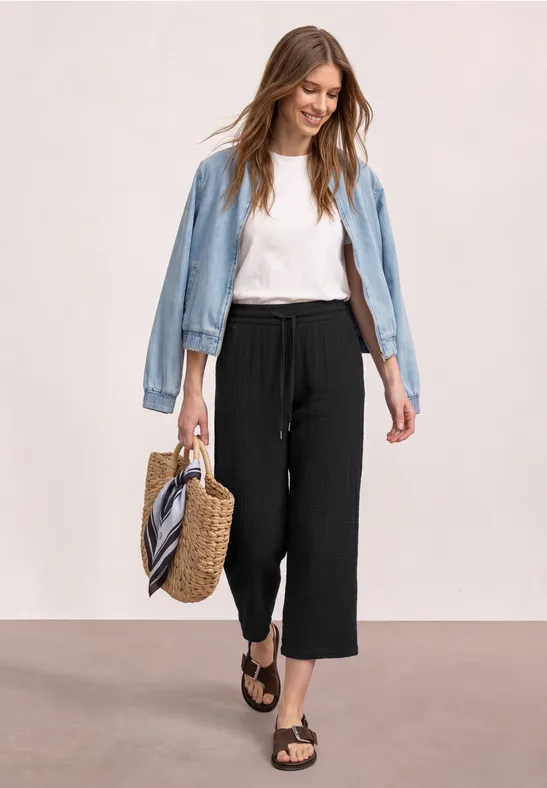 Frau in weißem Shirt, schwarzen Culottes, Jeansjacke, Basttasche und Sandalen vor hellem Hintergrund.