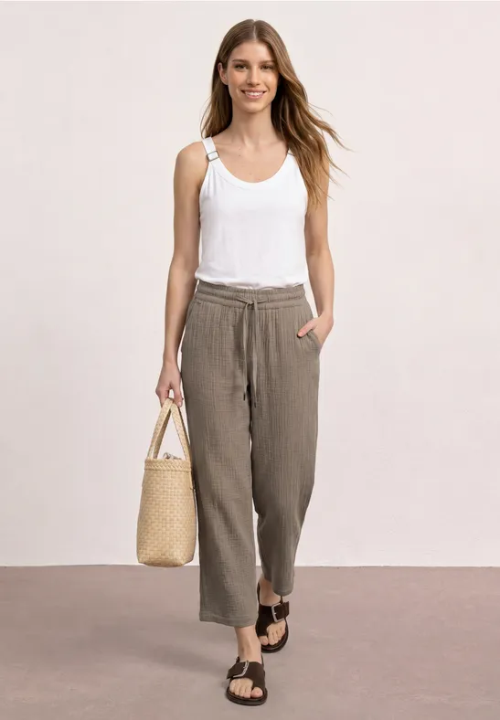 Vrouw met wit hemdje, losse bruine broek, sandalen en rieten tas, staand voor een neutrale achtergrond.